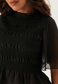 Sort kjole med en smocket bodice med teksturerede volanger, gennemsigtige korte ærmer og en høj halsudskæring, lavet af letvægtsstof.