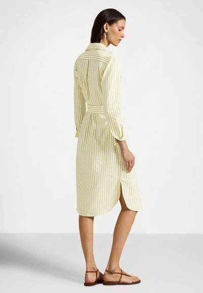 Kvinne iført en gul og hvit kjole med vertikale striper, langermet, med belte i midjen og brune flate sandaler, stående i profil mot en hvit bakgrunn.