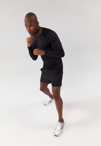 Black long-sleeve athletic top and black shorts worn by a man. He wears silver running shoes and is in a fighting stance. Plain background.
