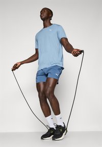 T-shirt de sport bleu clair, shorts bleus, chaussures de course noires. Corde à sauter tenue dans les mains. Design simple avec des accents de logo sur les vêtements.