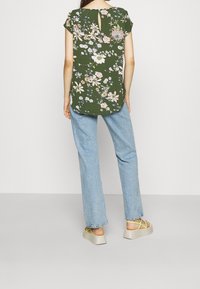 Groen bloemen topje met korte mouwen, een open rug met keyhole detail, gecombineerd met lichtblauwe spijkerbroek en lichte schoenen met gele accenten.