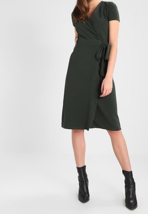 Femme portant une robe portefeuille vert foncé longueur genou avec des manches courtes et des bottines noires, debout devant un fond uni.