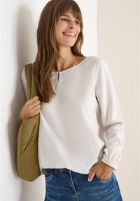 Blusa grigia chiaro a maniche lunghe con scollatura rotonda, caratterizzata da un piccolo dettaglio a forma di chiave. Tessuto texturizzato e polsini elasticizzati. Abbinata a una borsa verde.