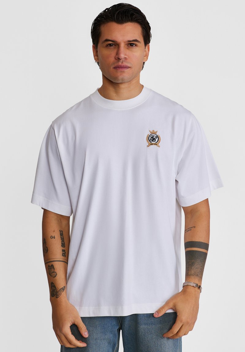 Homme portant un T-shirt blanc oversize avec un petit blason brodé sur le côté gauche de la poitrine, un jean bleu et des tatouages visibles sur les deux bras.