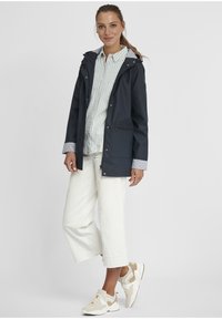 Veste imperméable navy avec fermeture éclair et pression, dotée de deux poches avant et de poignets rayés. Associée à un pantalon blanc coupe courte et des baskets claires.