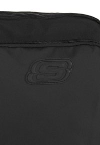 Bolsa de tela negra con una textura suave, que presenta un prominente logo negro en relieve. El zipper y las costuras están alineados de manera ordenada.