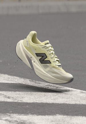 Chaussure de course New Balance jaune clair avec logo noir apparaissant en l'air au-dessus de l'asphalte avec des marquages routiers blancs.