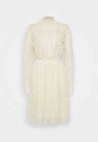 Lace och prickig krämfärgad klänning med hög krage, lång ärm, volangdetaljer och flerlagers kjol, som har genomskinligt och strukturerat tyg.