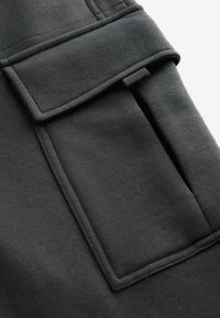 Schwarze Stofftasche mit rechteckigem Deckel, genähten Details und strukturierter Oberfläche, Teil eines Kleidungsstücks, das Funktionalität und Design betont.