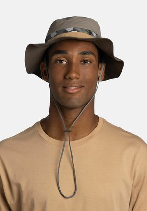 Joven con un sombrero tipo pescador beige de ala ancha con correa para la barbilla y una camiseta beige claro de cuello redondo, sonriendo suavemente.