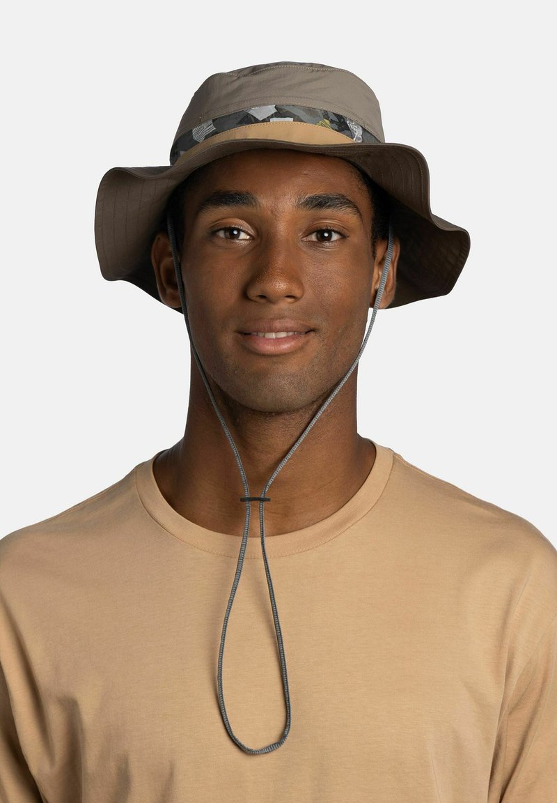 Joven con un sombrero tipo pescador beige de ala ancha con correa para la barbilla y una camiseta beige claro de cuello redondo, sonriendo suavemente.
