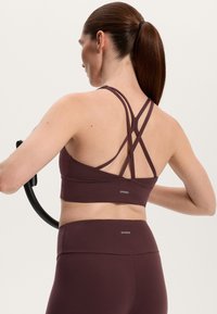 Burgundyröd sportbh med korsade remmar, mjukt elastiskt material och logotypdetalj vid basen, ihop med matchande högmidjade leggings.