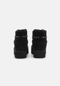Stivali ankle in suede nero con fodera in pelliccia sintetica, suole basse e lacci bianchi e neri a contrasto. Design minimalista, nessun logo visibile.