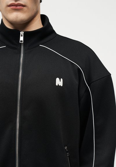 Άνδρας με μαύρο μπουφάν με φερμουάρ και λευκή λεπτομέρεια, με μικρό λευκό λογότυπο "N" στο αριστερό στήθος, μπροστά σε απλό φόντο.