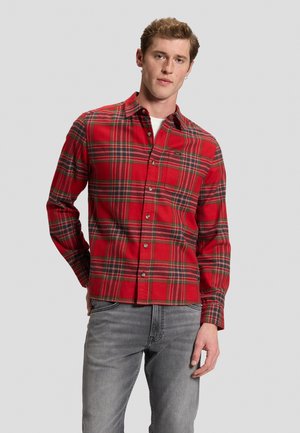 Jeune homme portant une chemise à carreaux rouge et verte avec un jean gris, debout avec une main derrière le dos sur un fond uni.