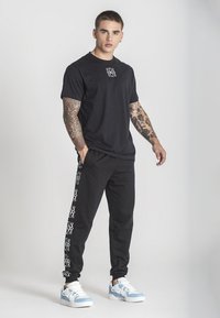 Svart bomull t-shirt med en liten vit grafik på bröstet, tillsammans med svarta joggers som har vita mönster längs sidorna. Blå och vita sneakers.