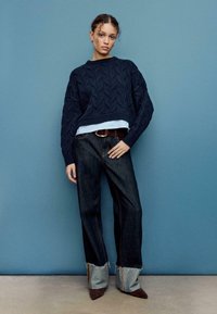 Maglione lavorato a maglia blu navy con motivo a trecce indossato sopra una maglietta azzurro chiaro, abbinato a jeans a gamba larga in denim scuro e polsini grigi. Scarpe marroni con punta appuntita.