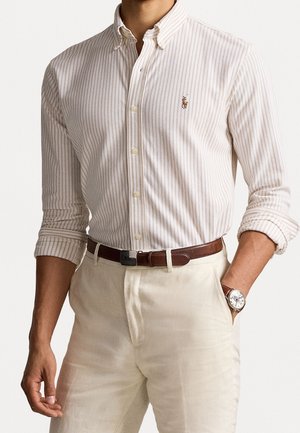 Homme portant une chemise beige rayée à boutons avec manches retroussées, pantalon beige, ceinture marron et montre-bracelet avec bracelet en cuir.