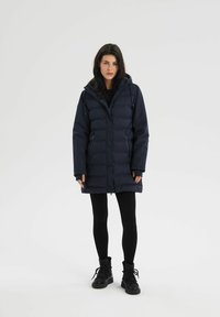 Granatowa kurtka puchowa o długim kroju, z kapturem, zamkiem błyskawicznym i kieszeniami po bokach, w połączeniu z czarnymi legginsami i czarnymi butami za kostkę na sznurowadła.