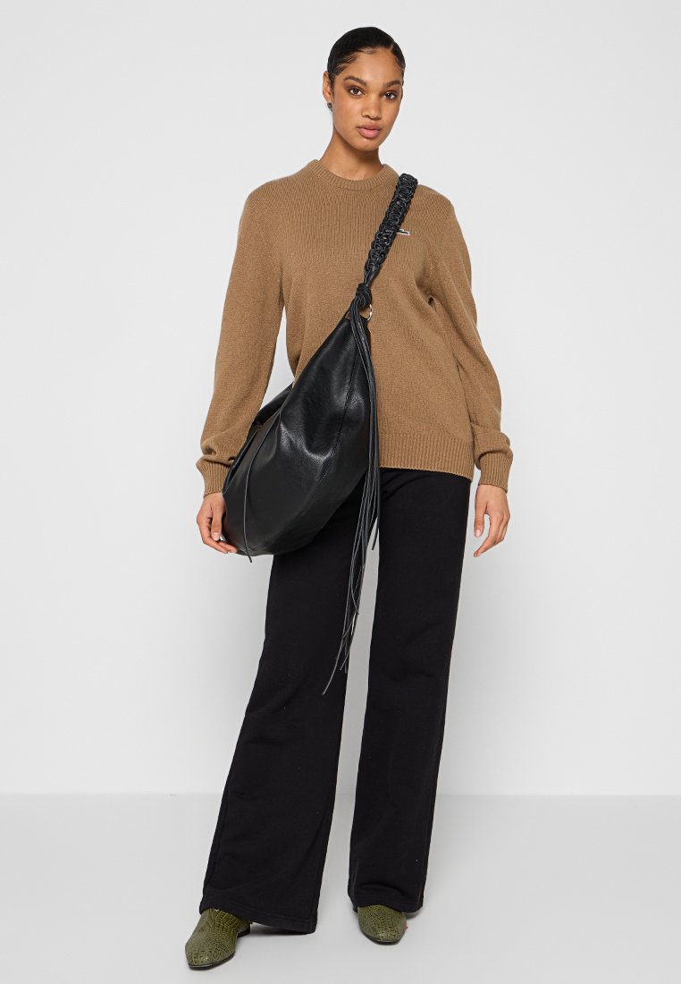 Borsa hobo in pelle nera con manico intrecciato, caratterizzata da dettagli con frange. Il modello indossa un pullover beige e pantaloni neri a gamba larga. Calzature verdi.