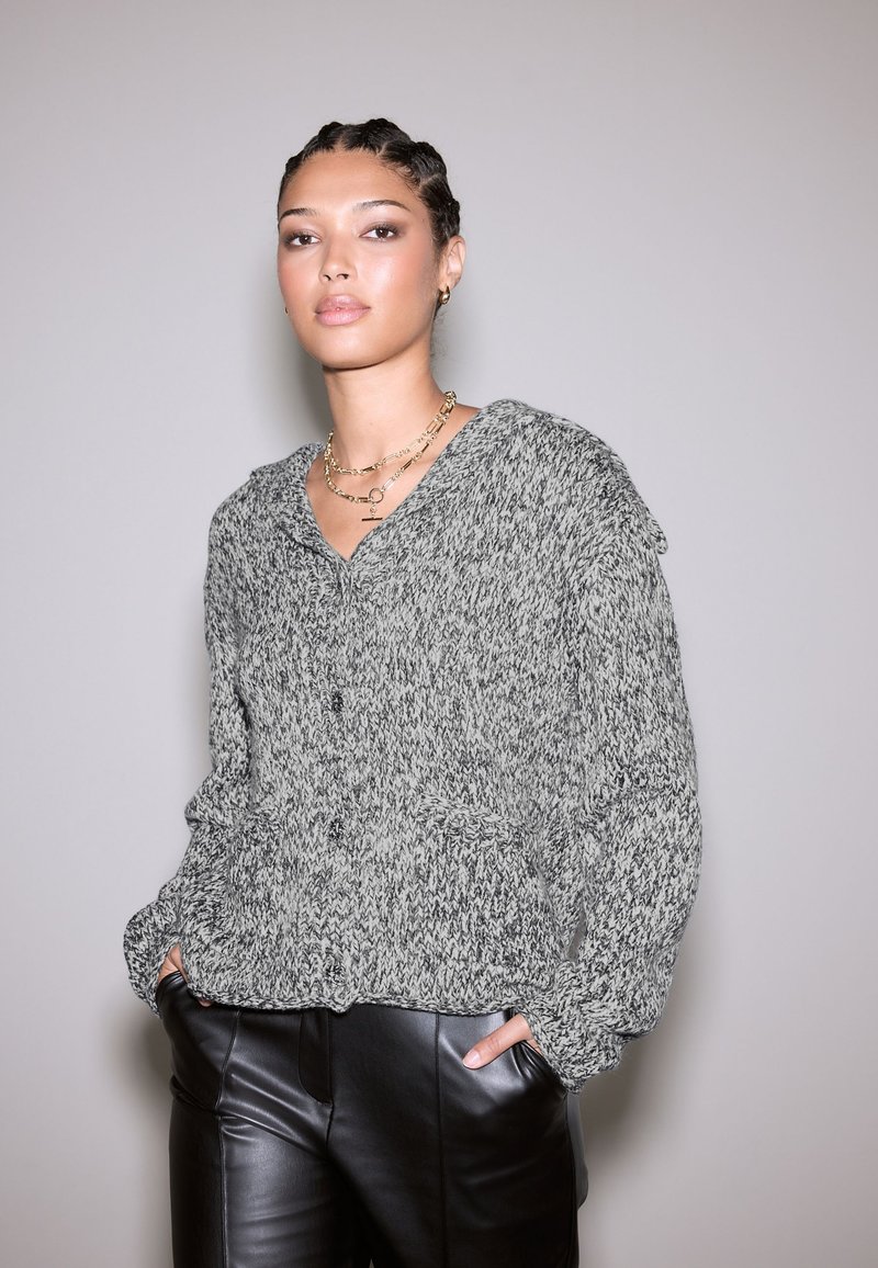 Sivi i crni pleteni kardigan s prednjim kopčanjem na gumbe, V-izrezom i bočnim džepovima. Nošen preko crnih kožnih hlača.