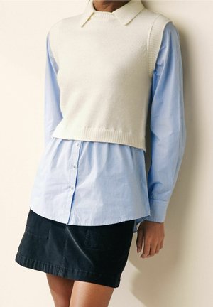 Personne portant un pull sans manches blanc en laine par-dessus une chemise bleue rayée à boutons et une jupe noire, se tenant devant un mur clair.