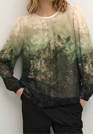 Femme portant une blouse transparente à manches longues avec un dégradé floral vert et beige, associée à un pantalon noir à fines rayures, les mains dans les poches.