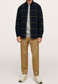Camicia di flanella a quadri blu navy e nera, con colletto button-down, indossata sopra un maglione grigio chiaro. I pantaloni color sabbia e le sneakers beige completano il look.