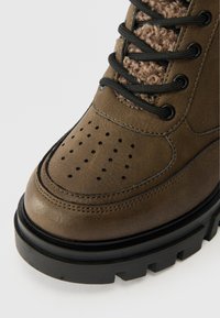 Hnědá kožená bota s texturovaným fleecovým akcentem, perforovanou špičkou, černými tkaničkami a silnou černou platformovou podrážkou. Obsahuje robustní mřížkové otvory.