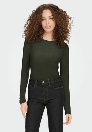 Olijfgroene top met lange mouwen in een textuurstof, gecombineerd met hoge donkere jeans met gouden stiksels en een knoopsluiting.