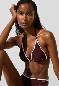 Pruun bikiinide komplekt, mis sisaldab kolmnurkset braletti valge lainelise äärisega ja vastavaid aluspükse, millel on reguleeritavad seotud detailid ja peene logo.