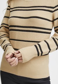 Pull en tricot beige avec des rayures horizontales noires, tissu texturé et poignets côtelés. Possède des ouvertures pour les pouces dans le design des manches.