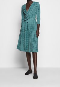 Robe portefeuille sarcelle à manches trois-quarts, présentant un motif tourbillonnant, taille ceinturée et jupe évasée. Portée avec des mocassins noirs.