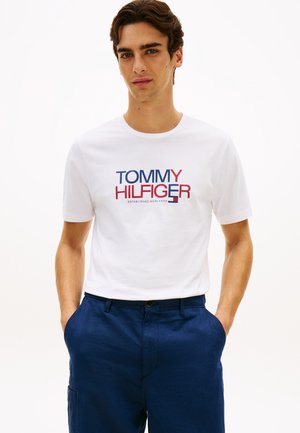 Mann iført hvit Tommy Hilfiger-logo t-skjorte og blå bukser, stående med hendene i lommene foran ensfarget bakgrunn.