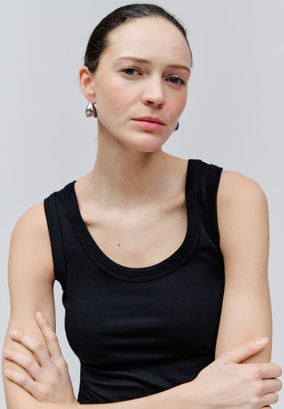 Jonge vrouw met bleke huid en blauwe ogen, die zilveren oorbellen draagt en een zwart hemdje, met gekruiste armen tegen een effen grijze achtergrond.