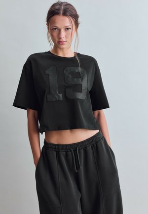 Junge Frau trägt ein schwarzes, bauchfreies T-Shirt mit der Nummer 19 und passende schwarze Jogginghosen, steht vor einem schlichten hellgrauen Hintergrund.