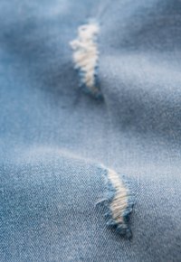 Tissu en denim bleu clair avec des fils blancs visibles dans les zones effilochées. La texture de la surface est lisse avec de légères variations de couleur.