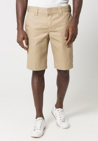 Beige shorts tillverkade av lättviktsmaterial, med rak passform, platt front och sidofickor, i kombination med vita sneakers.