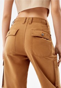 Cargo-byxor i en varm brun färg, tillverkade av slitstarkt tyg, med två bakfickor med lock och metallknappar för stängning.