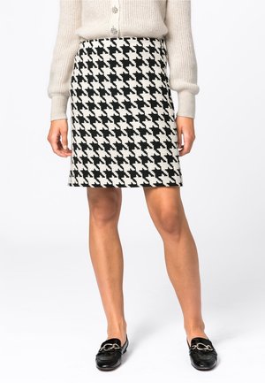 Woman wearing a black and white houndstooth skirt, beige buttoned sweater, and black slip-on loafers with metal detailing.