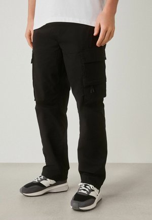 Person trägt schwarze Cargo-Hosen mit großen Seitentaschen und schwarz-weißen Sneakers und steht auf einem hellgrauen Boden.