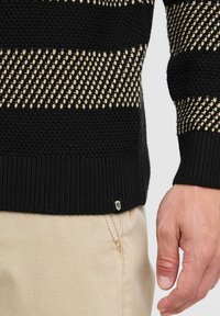 Black knitted sweater featuring horizontal cream stripes, textured patterns, and a ribbed hem with a small logo tag. Paired with beige pants.