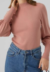 Maglione lavorato a maglia rosa chiaro con un collo alto e maniche lunghe a sbuffo, abbinato a pantaloncini in denim blu a vita alta.