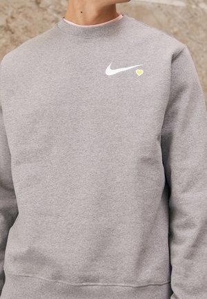 Sweatshirt gris en mélange de coton, arborant un logo Nike blanc et un petit accent en forme de cœur jaune sur la poitrine. Poignets et ourlet côtelés.
