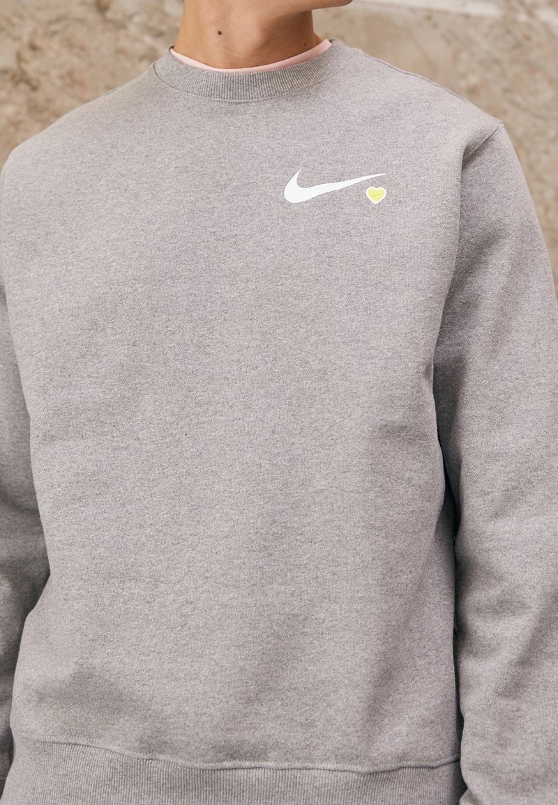 Grå tygtröja i bomullsmix med en vit Nike-logotyp och en liten gul hjärtaccent på bröstet. Ribbad mudd och nederkant.