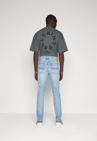 Camiseta de algodón gris con un gran logotipo negro en la parte posterior, combinada con jeans de mezclilla azules claros y zapatillas grises con acentos a rayas.