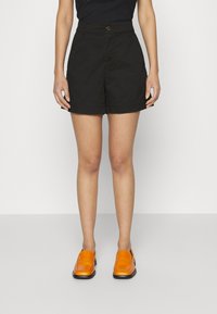 Černé bavlněné šortky s vysokým pasem, předním zapínáním na knoflíky a bočními kapsami; nošeny s jasně oranžovými nazouvacími botami.