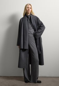 Mujer rubia que lleva un abrigo de lana gris oscuro en capas, bufanda, suéter de cuello alto y pantalones de pierna ancha, de pie contra una pared clara y sencilla.