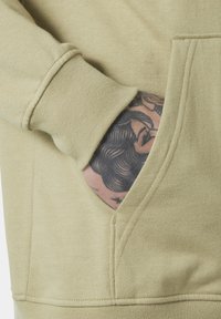 Sudadera de color beige claro con un bolsillo frontal y puños acanalados. La textura parece suave, con líneas de costura visibles y un tatuaje visible en el bolsillo.
