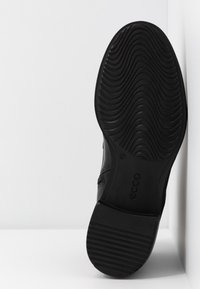Suela de goma negra de un zapato que presenta una textura ondulada y un diseño acanalado. El zapato tiene un contorno superior suave y lleva el nombre de la marca "ECCO."